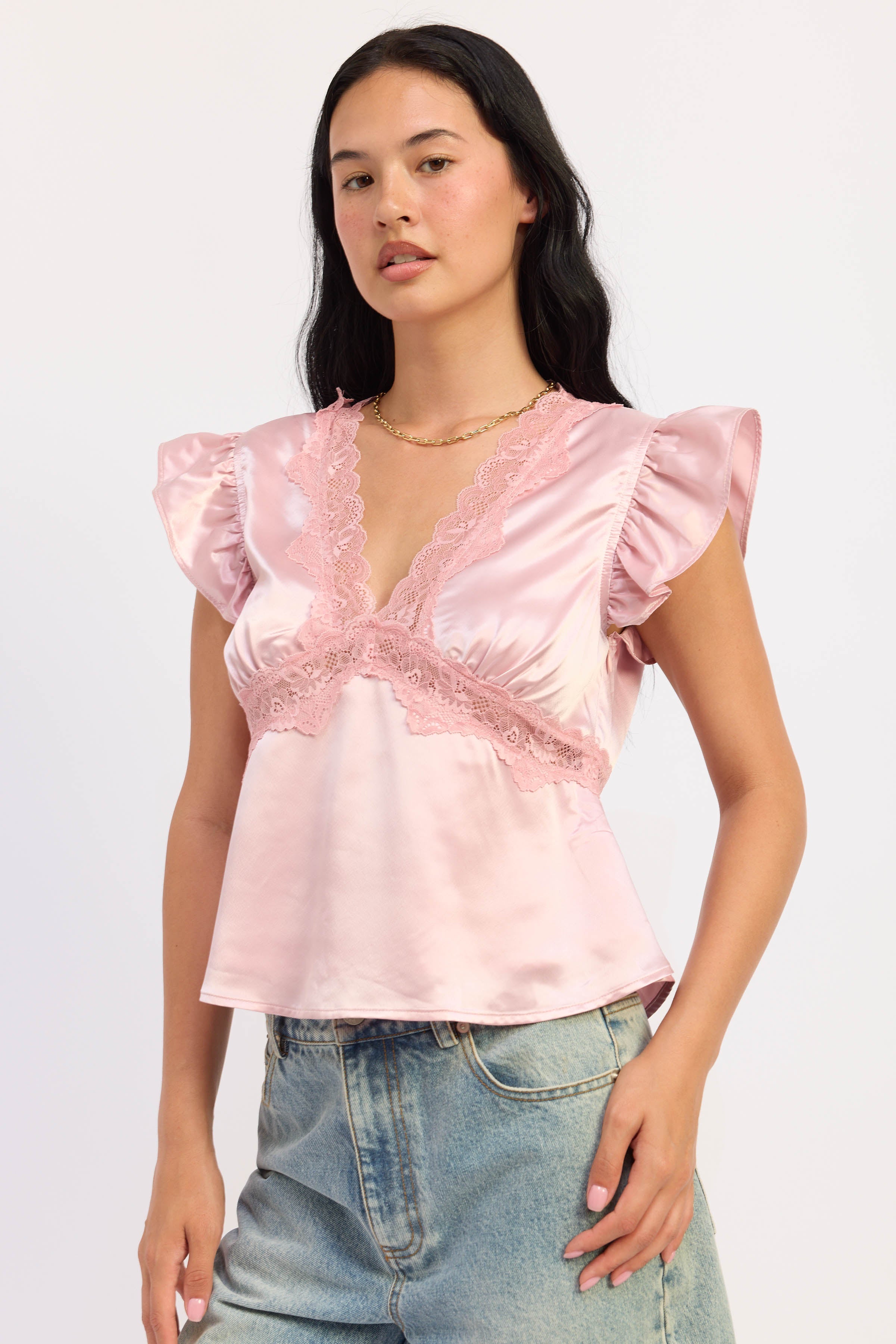 A woman modeling a light pink satin top with ruffled cap sleeves, a lace-trimmed V-neckline, and a gathered peplum waist, paired with light-wash denim jeans against a plain white studio background.