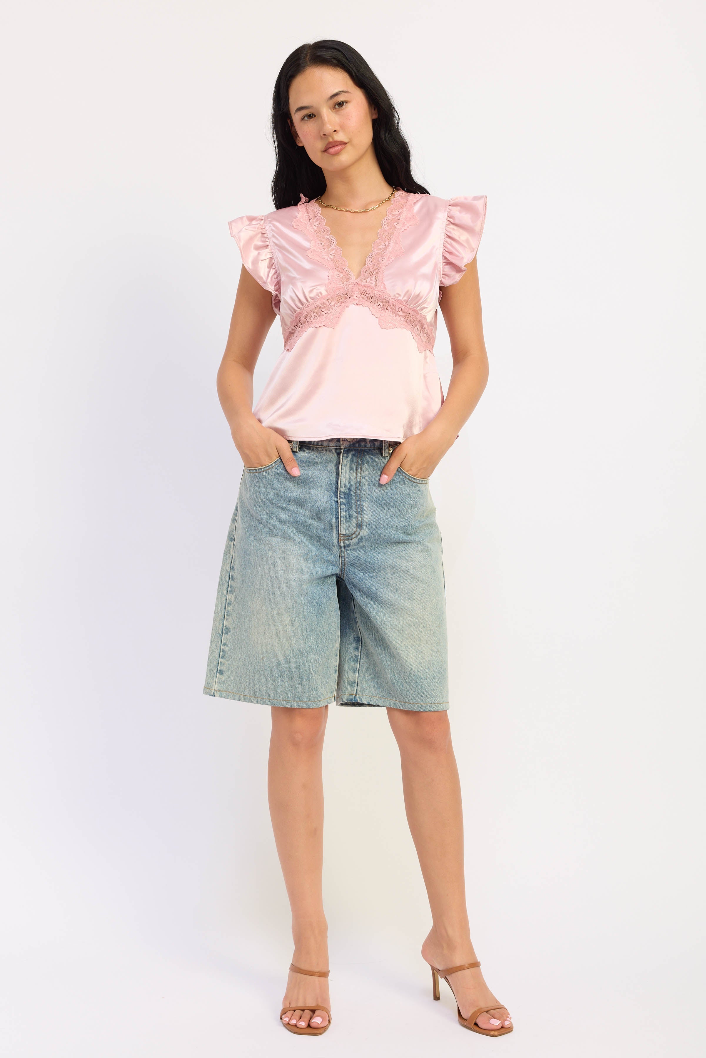 A woman modeling a light pink satin ruffled top paired with light-wash denim shorts and brown heeled sandals, standing against a plain white studio background.