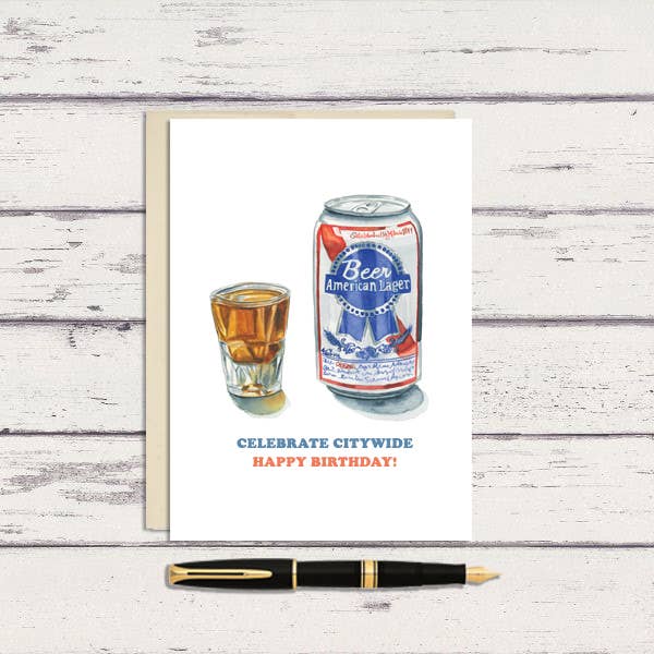 A greeting card with a watercolor illustration of a beer can and a glass, alongside a pen. The card is placed on a wooden surface.