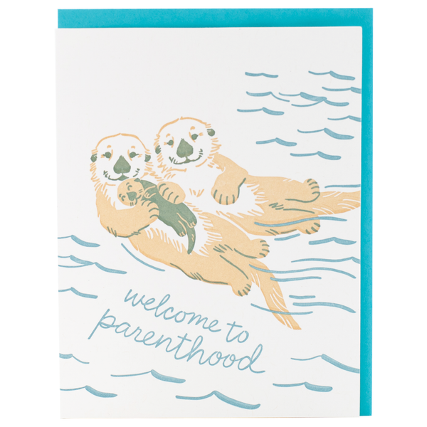 A greeting card with an illustration of two otters floating in water, with one otter holding a baby otter. The text on the card reads 'welcome to parenthood'.