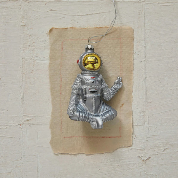 A hand-painted glass astronaut ornament with a peace sign and glitter, featuring a silver and gold finish, displayed on a textured wall.