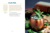 Copper mug with a drink garnished with mint leaves, surrounded by ingredients and a book on a wooden surface.