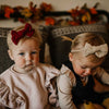 Two young children sitting on a couch with decorative bows in their hair, surrounded by autumn-themed decorations.