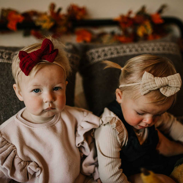 Two young children sitting on a couch with decorative bows in their hair, surrounded by autumn-themed decorations.