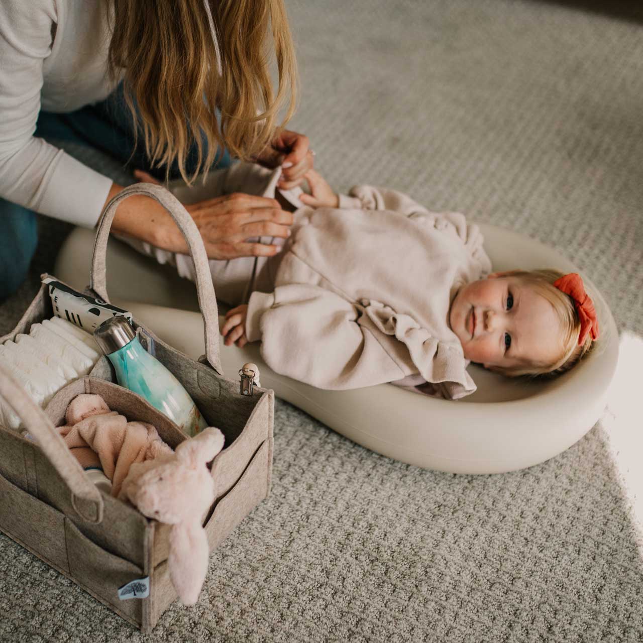 Baby lying in a white bassinet with a woman adjusting something near them, on a textured floor.