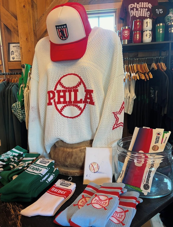 Mannequin wearing a white sweater with 'PHILA' text, red cap, and various sports-themed items on a table.