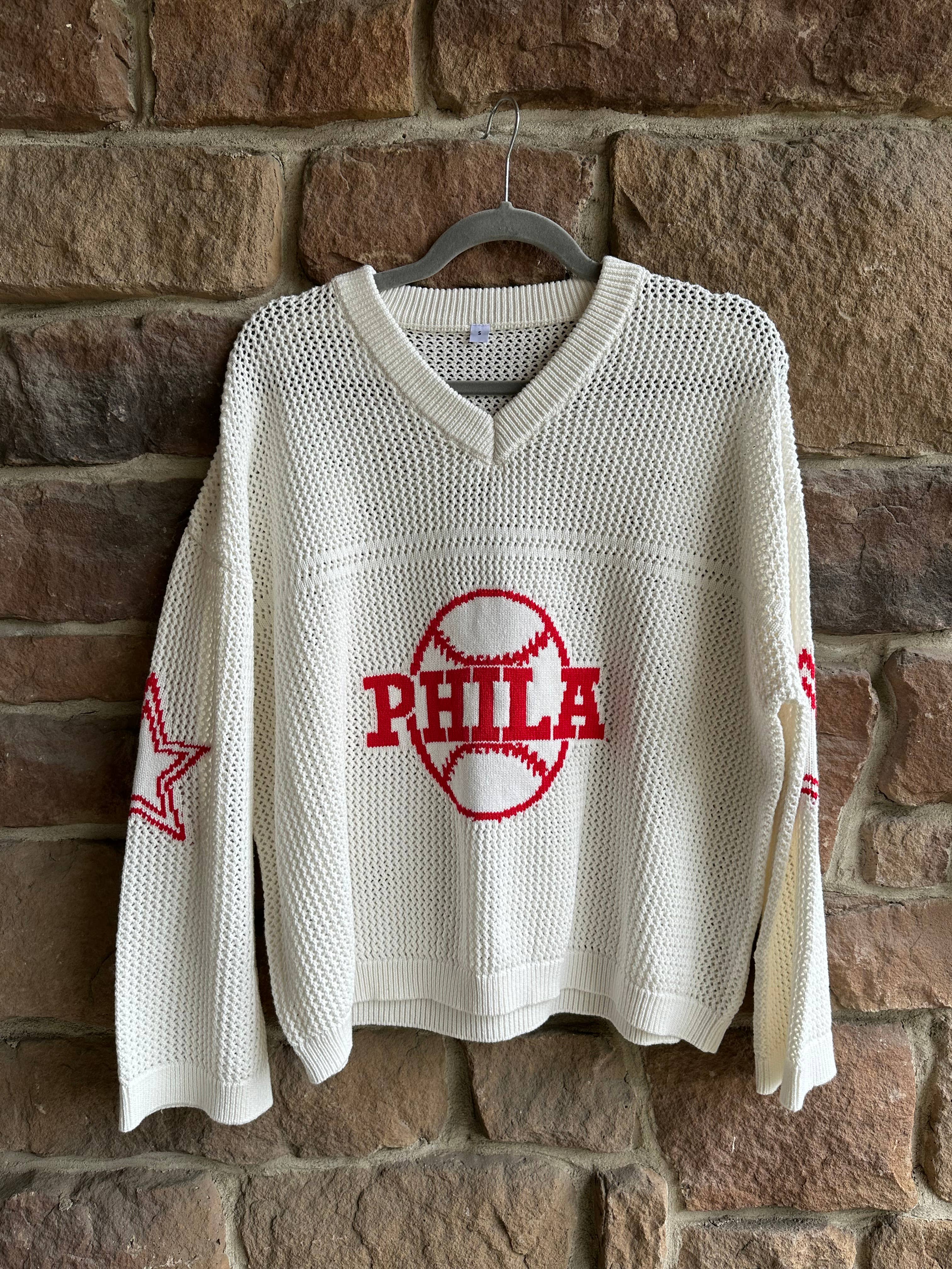 White sweater with red 'PHILA' text and star design on a hanger against a stone wall.