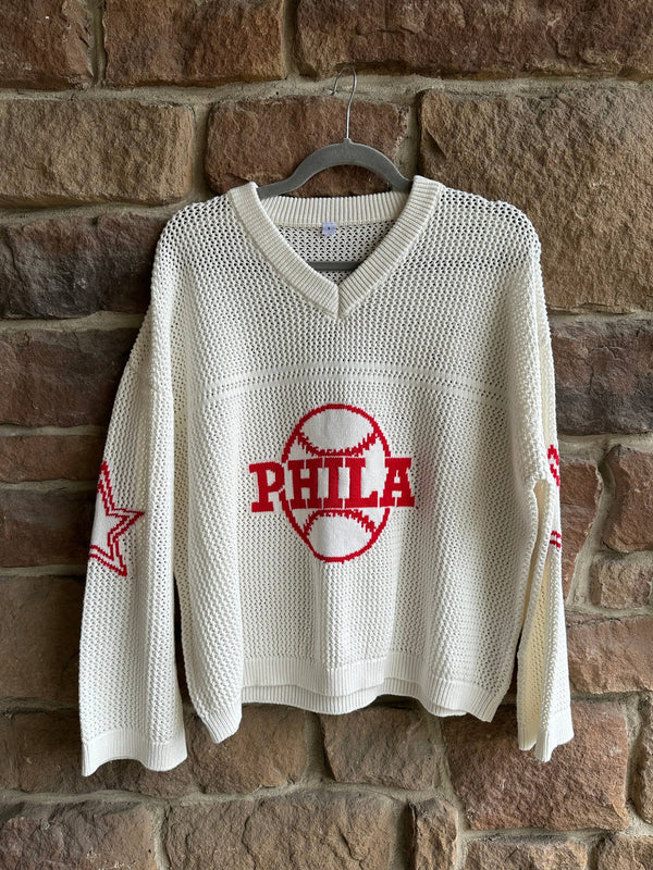 White sweater with red 'PHILA' text and star design on a hanger against a stone wall.