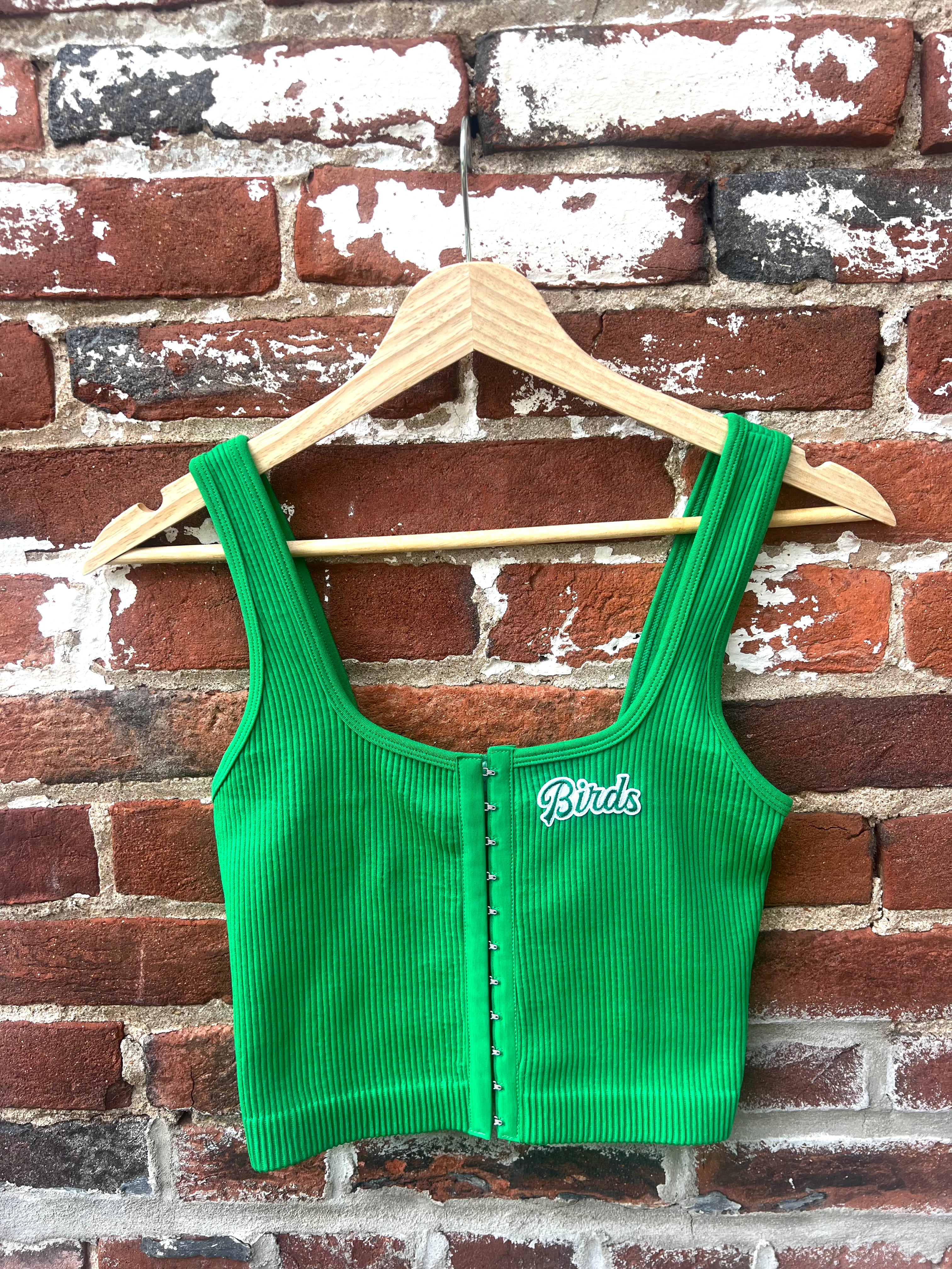 Green corduroy vest with 'Birds' logo on a hanger against a brick wall.