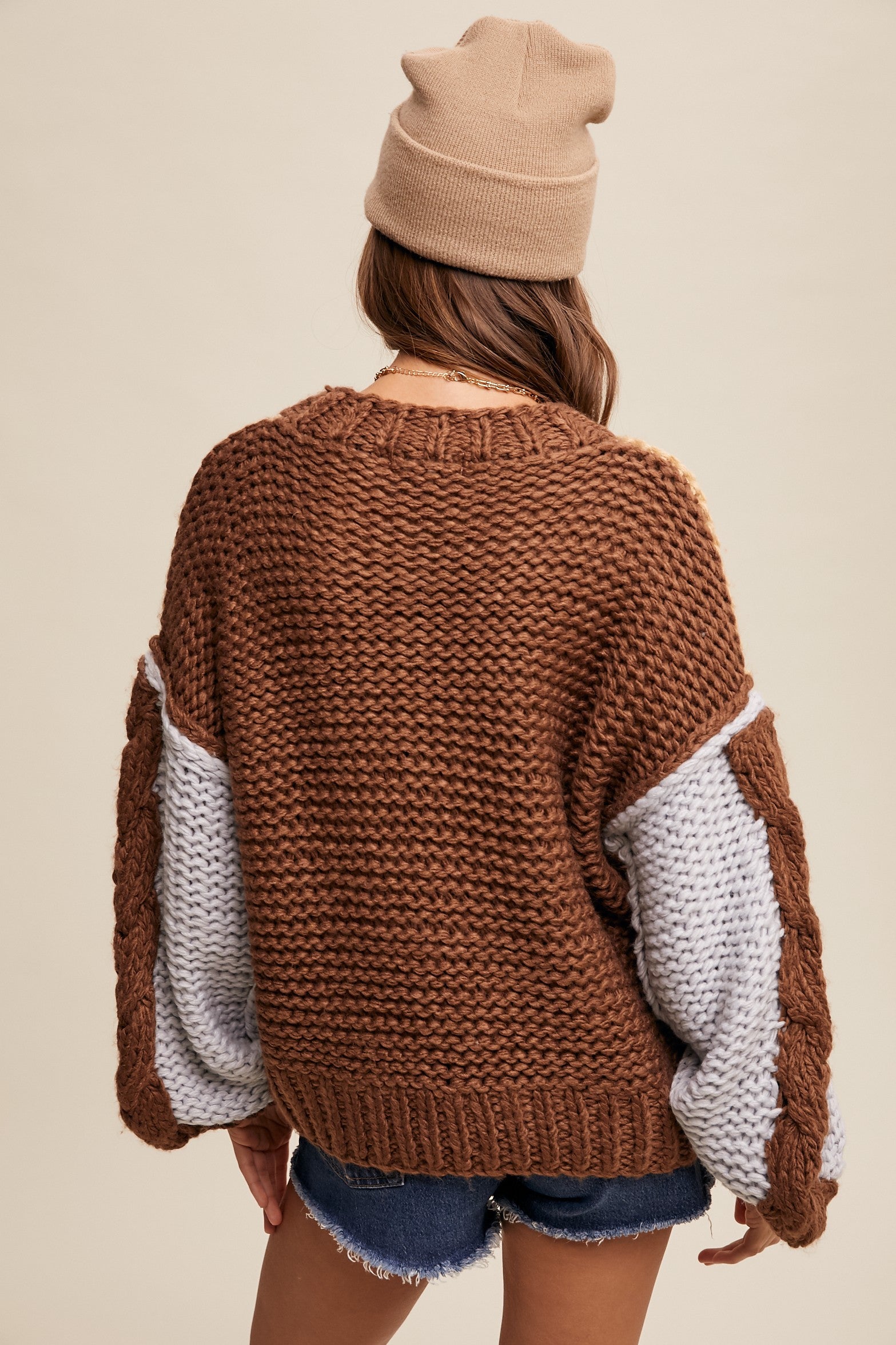 Person wearing a brown knitted sweater with white sleeves and a beige beanie on a beige background
