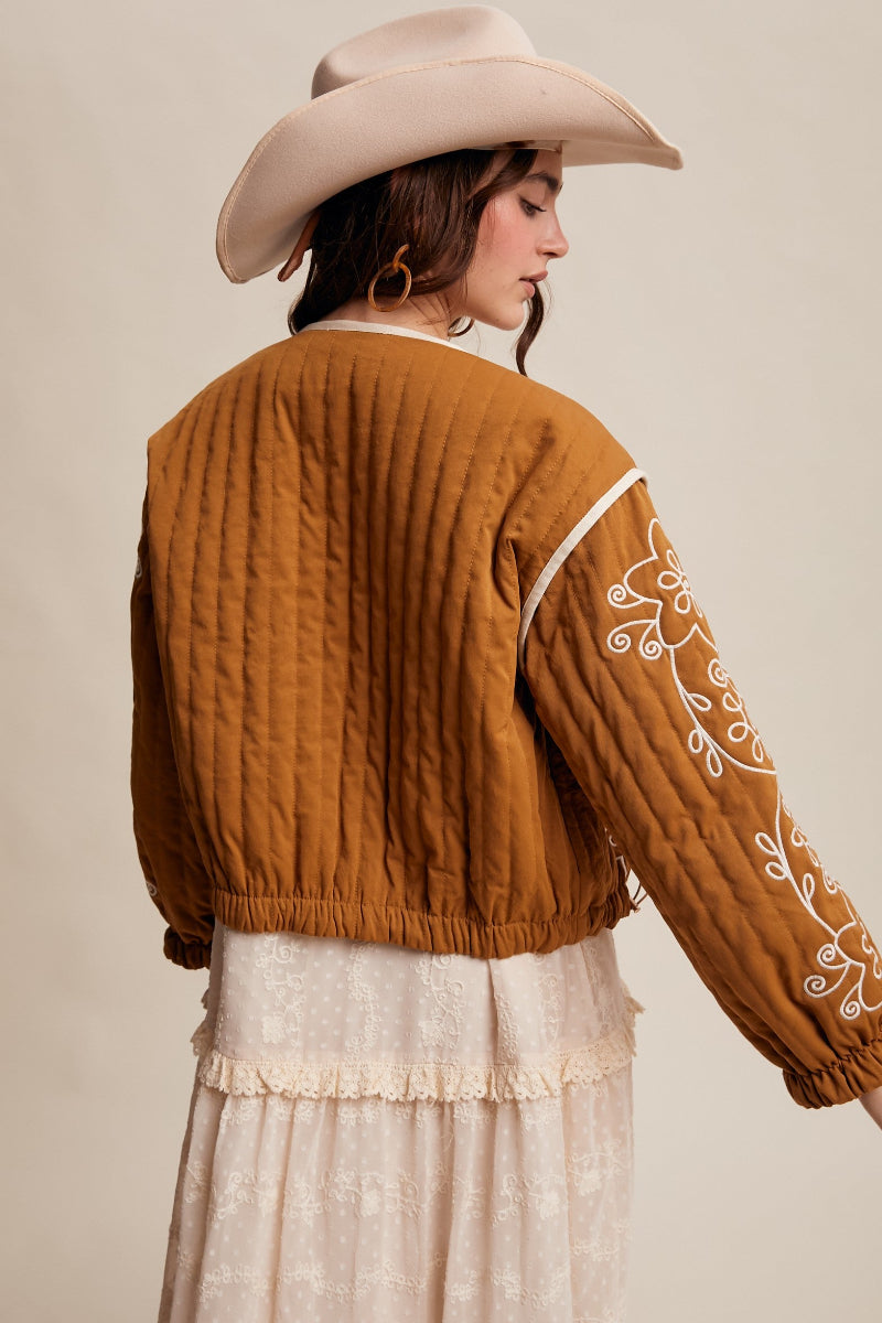 Woman wearing a brown quilted jacket with white embroidery and a beige cowboy hat on a beige background
