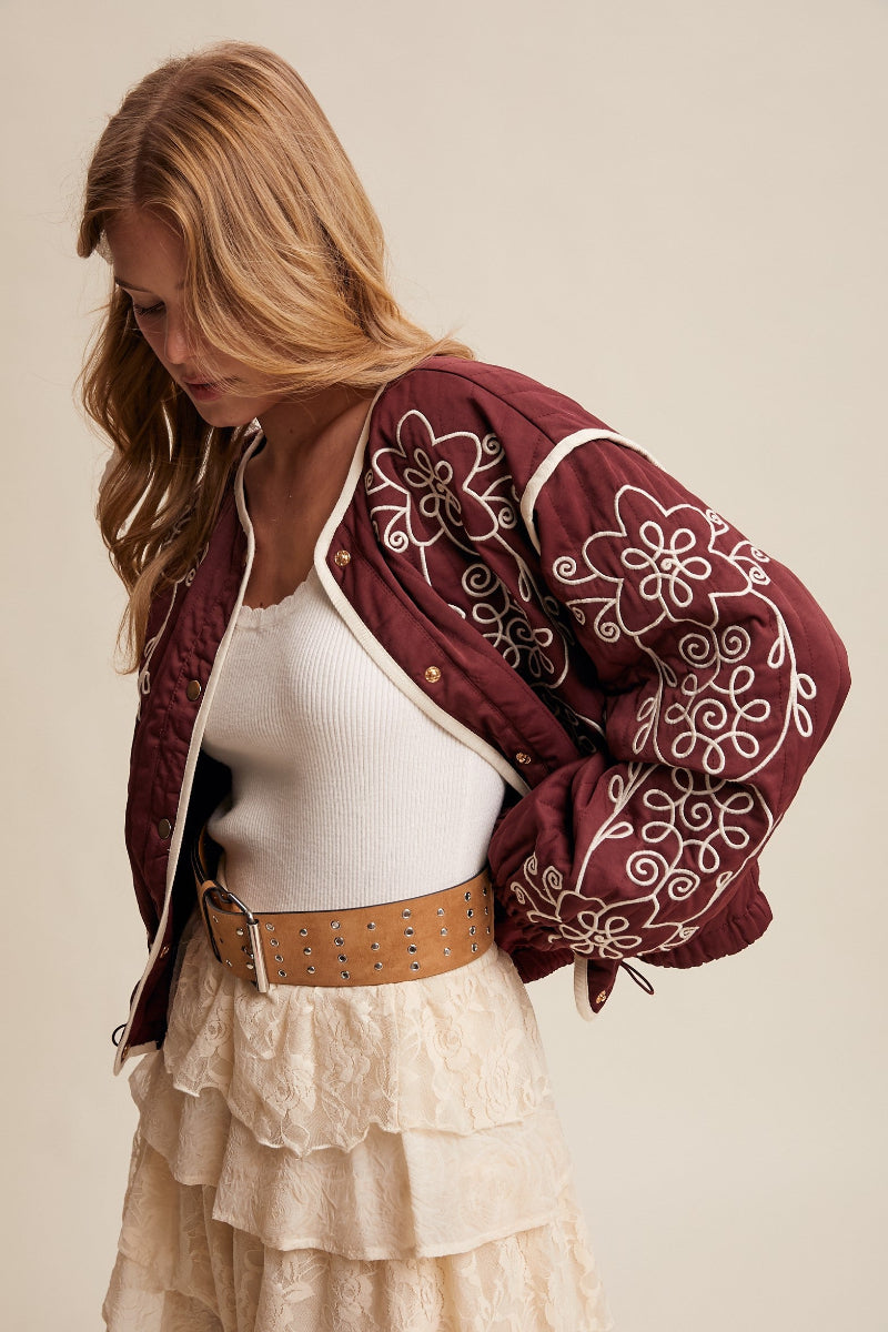 Woman wearing a burgundy embroidered jacket with white patterns, white top, and beige skirt on a beige background.
