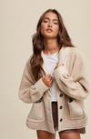 Woman wearing a beige cardigan over a white shirt and brown shorts on a plain background