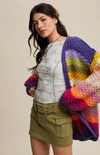 Woman wearing a colorful knitted cardigan over a white shirt and green shorts on a beige background