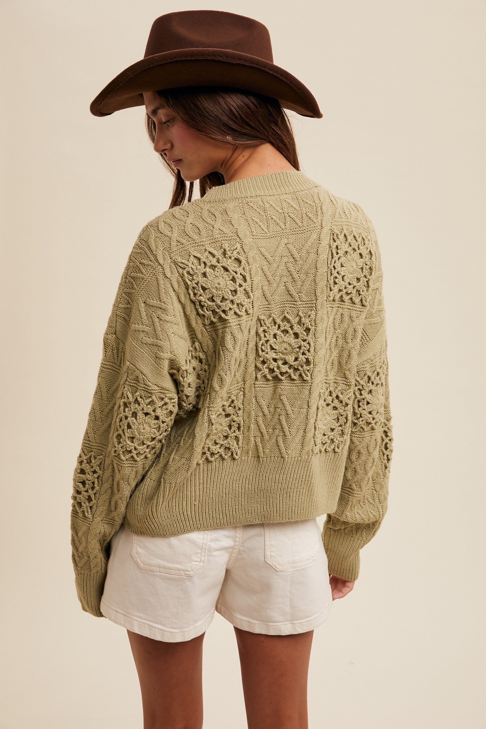 Person wearing a green knitted cardigan with lace details, white shorts, and a brown hat on a beige background.