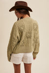 Person wearing a green knitted cardigan with lace details, white shorts, and a brown hat on a beige background.