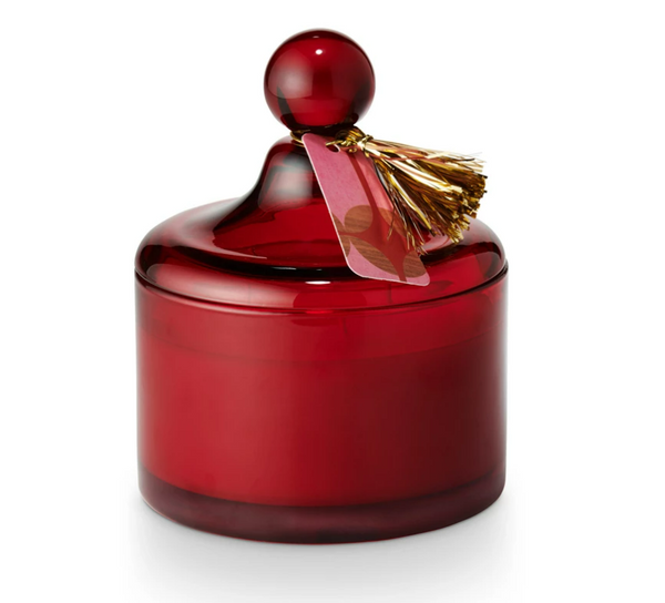 A red lidded glass candle with a Christmas-themed decoration on top.