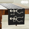 A black table runner with a pattern of white skeletons dancing, laid out on a wooden table.