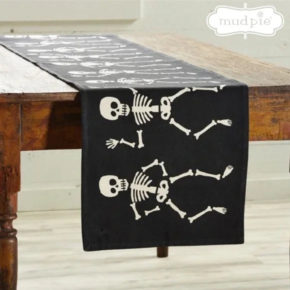 A black table runner with a pattern of white skeletons dancing, laid out on a wooden table.