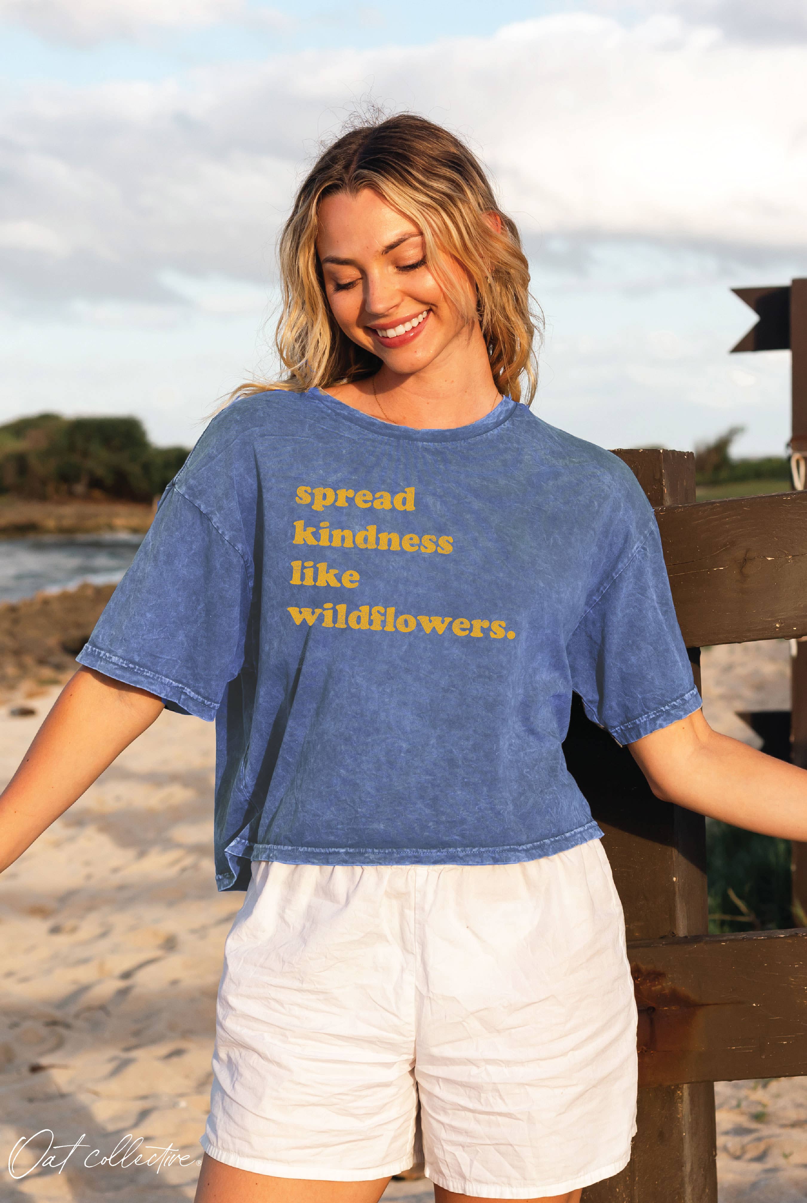Woman wearing a blue t-shirt with 'spread kindness like wildflowers' text on a beach.