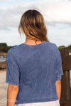 Person wearing a blue t-shirt sitting on a wooden bench by a body of water.