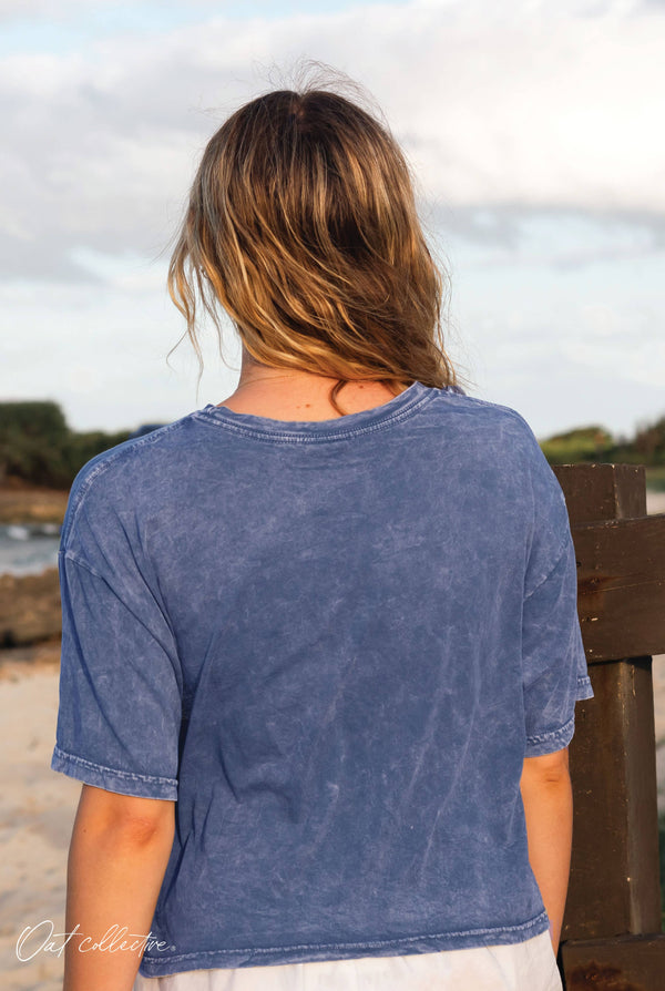 Person wearing a blue t-shirt sitting on a wooden bench by a body of water.