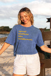 Woman wearing a blue t-shirt with 'spread kindness like wildflowers' text on a beach.
