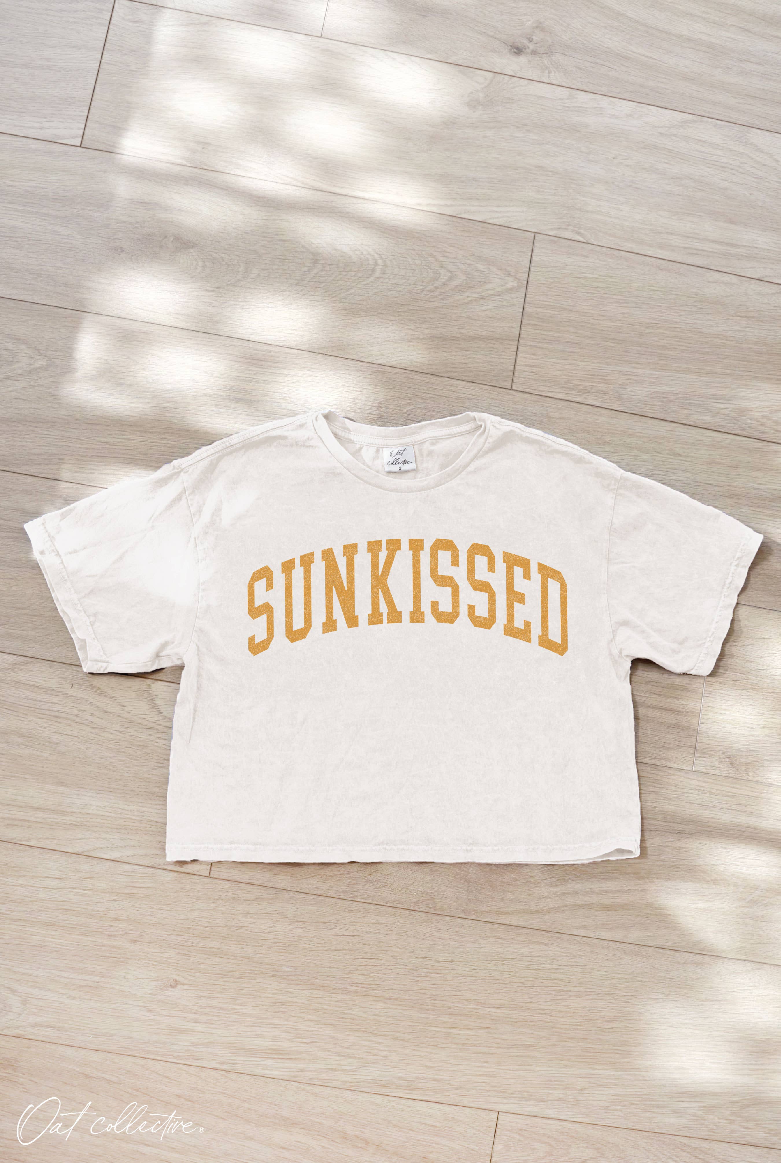 White t-shirt with 'SUNKISSED' text on a wooden floor