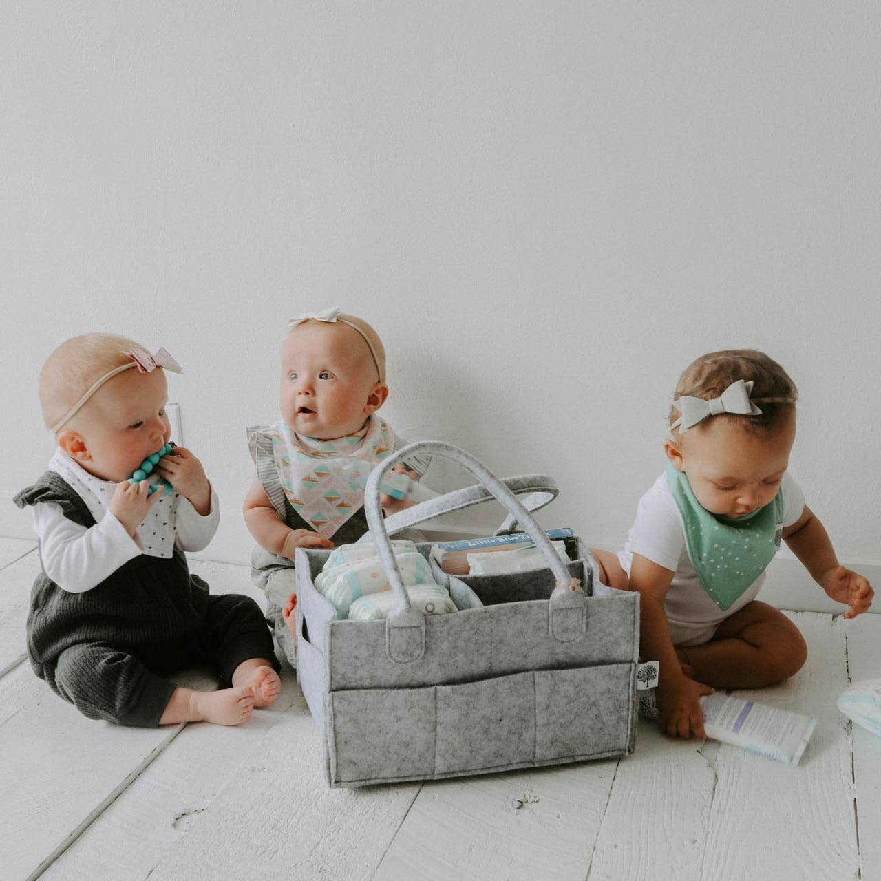 Three babies playing with a gray diaper bag on a white floor.