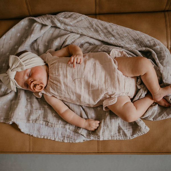 Newborn baby lying on a brown couch with a blanket, wearing a white headband.