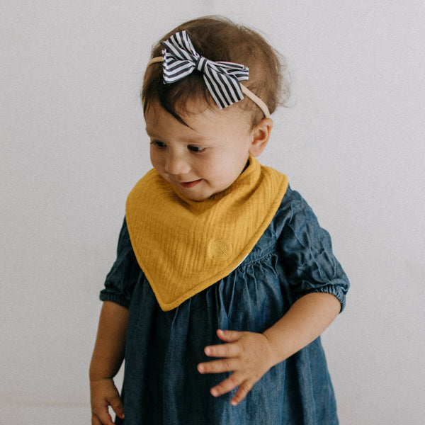 Child wearing a yellow bib and blue dress with a striped headband against a plain background