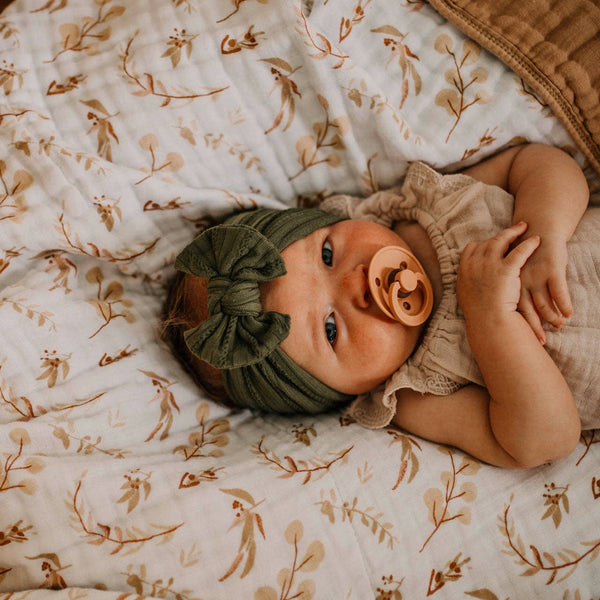 Baby lying on a floral blanket wearing a green headband and beige outfit with a pacifier.