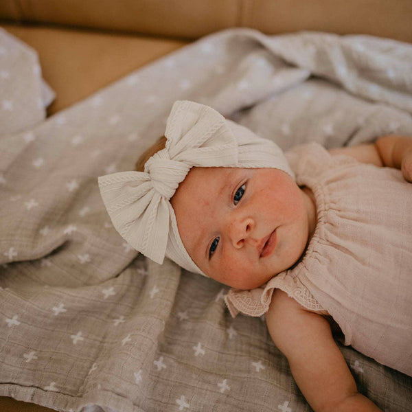 Baby wearing a beige bow headband lying on a patterned blanket.