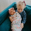Two children, one wrapped in a floral blanket and the other wearing a pink headband, sitting on a teal couch.