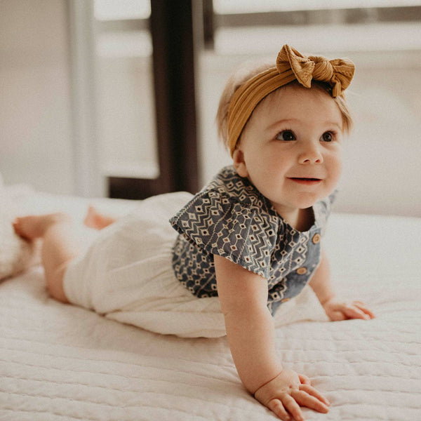 Baby crawling on a bed wearing a patterned romper and yellow headband.