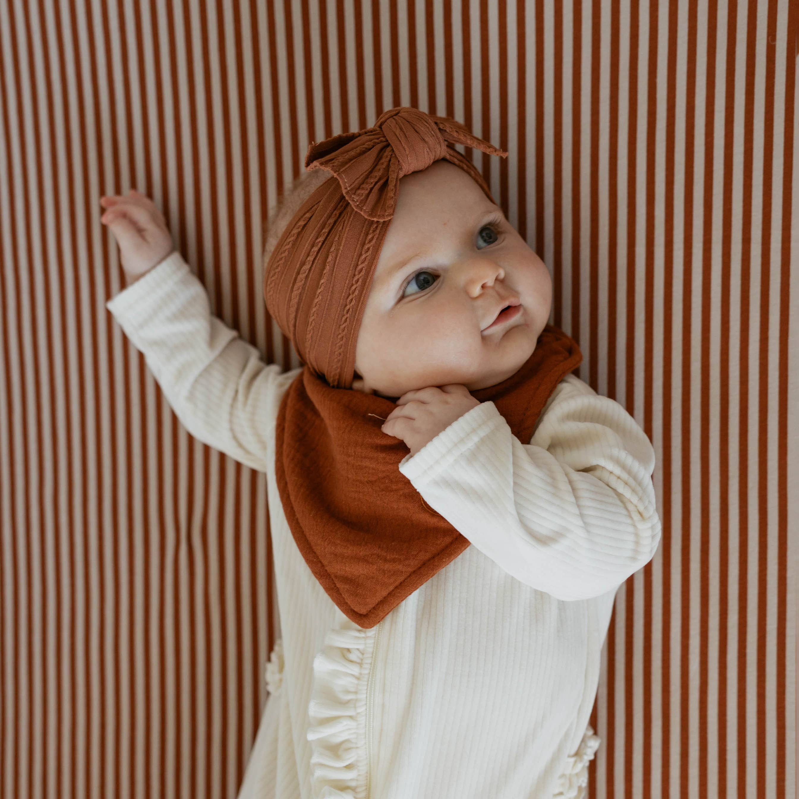 Baby wearing a rust-colored headband and bib against a striped background