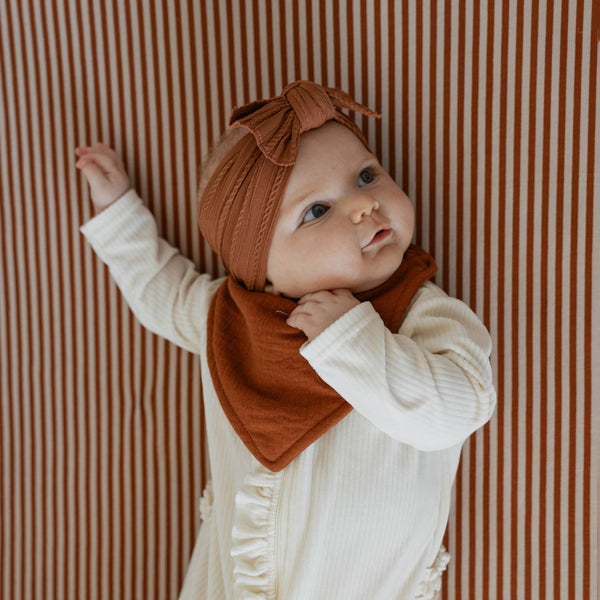 Baby wearing a rust-colored headband and bib against a striped background
