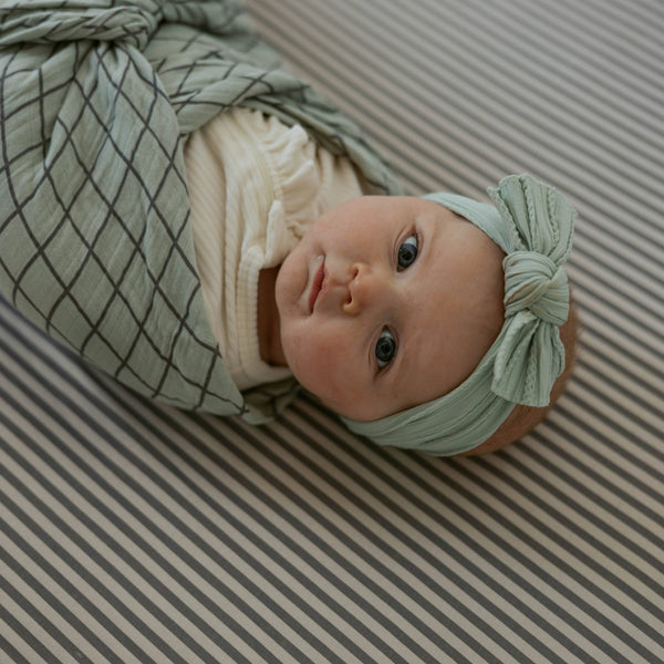 Newborn baby wrapped in a green swaddle with a matching headband on a striped background