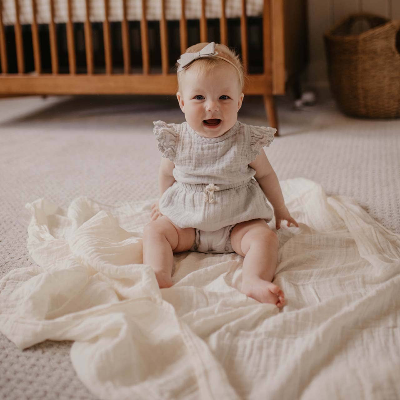 Baby sitting on a white blanket in a nursery with a wooden crib in the background.