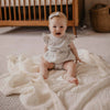 Baby sitting on a white blanket in a nursery with a wooden crib in the background.