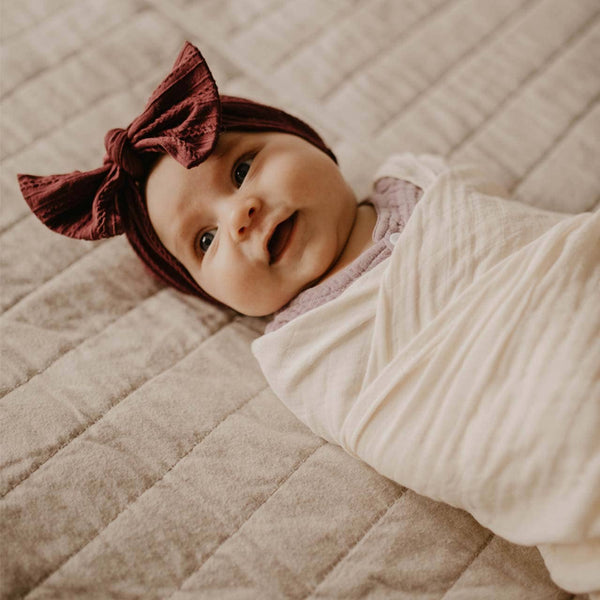 Baby wrapped in a white blanket with a maroon headband on a beige quilted surface