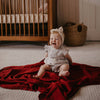Baby sitting on a red blanket in a nursery with a crib in the background