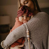 Woman holding a baby wrapped in a pink blanket with a headband, wearing a textured sweater.