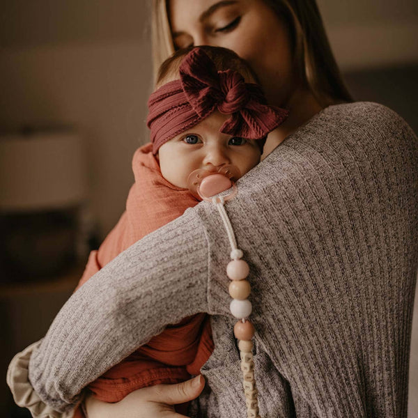Woman holding a baby wrapped in a pink blanket with a headband, wearing a textured sweater.