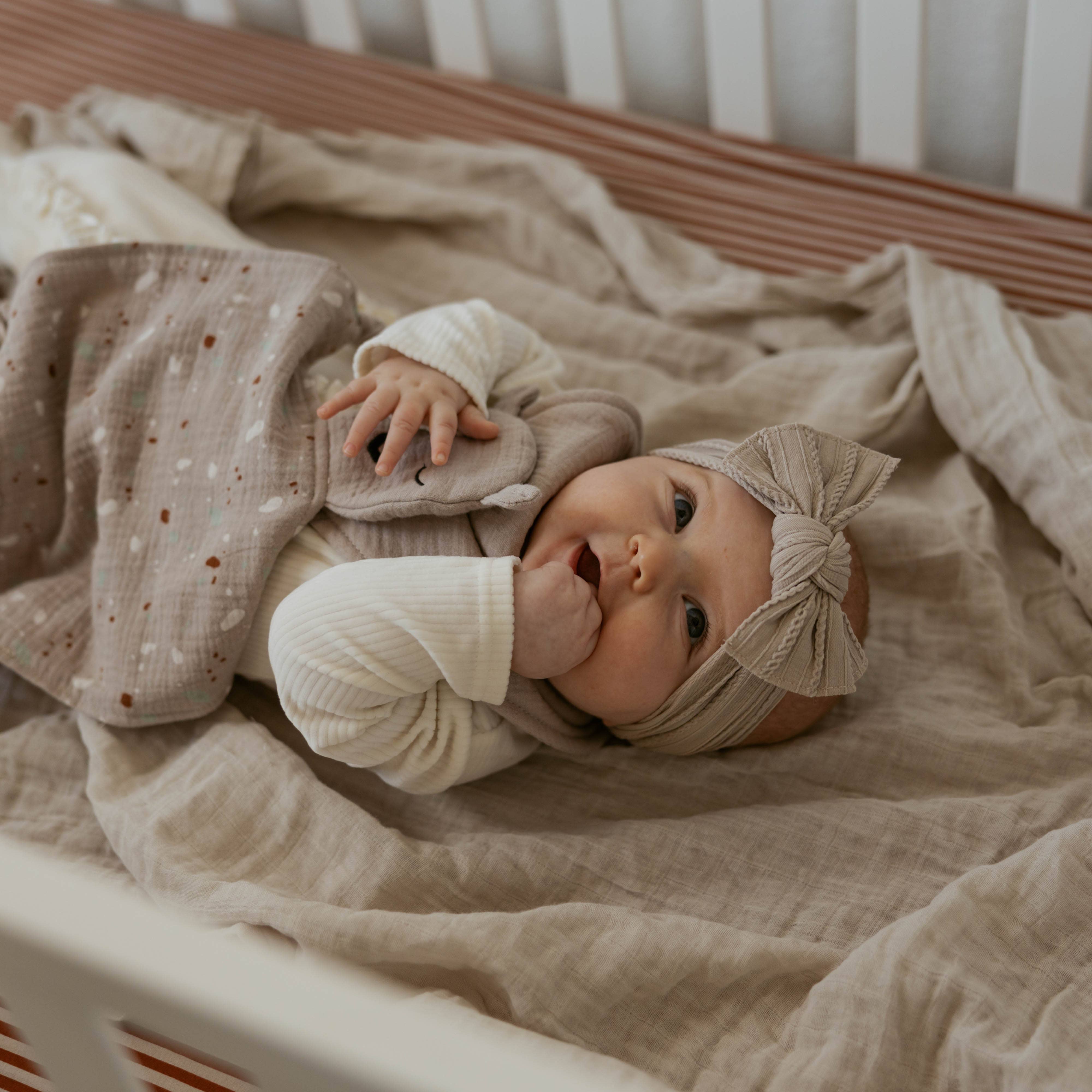Baby lying on a soft blanket in a crib wearing a beige headband.