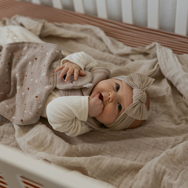 Baby lying on a soft blanket in a crib wearing a beige headband.