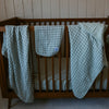 Wooden crib with green checkered blankets draped over it against a white paneled wall.