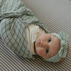 Newborn baby wrapped in a green swaddle with a matching headband on a striped background