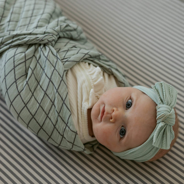 Newborn baby wrapped in a green swaddle with a matching headband on a striped background