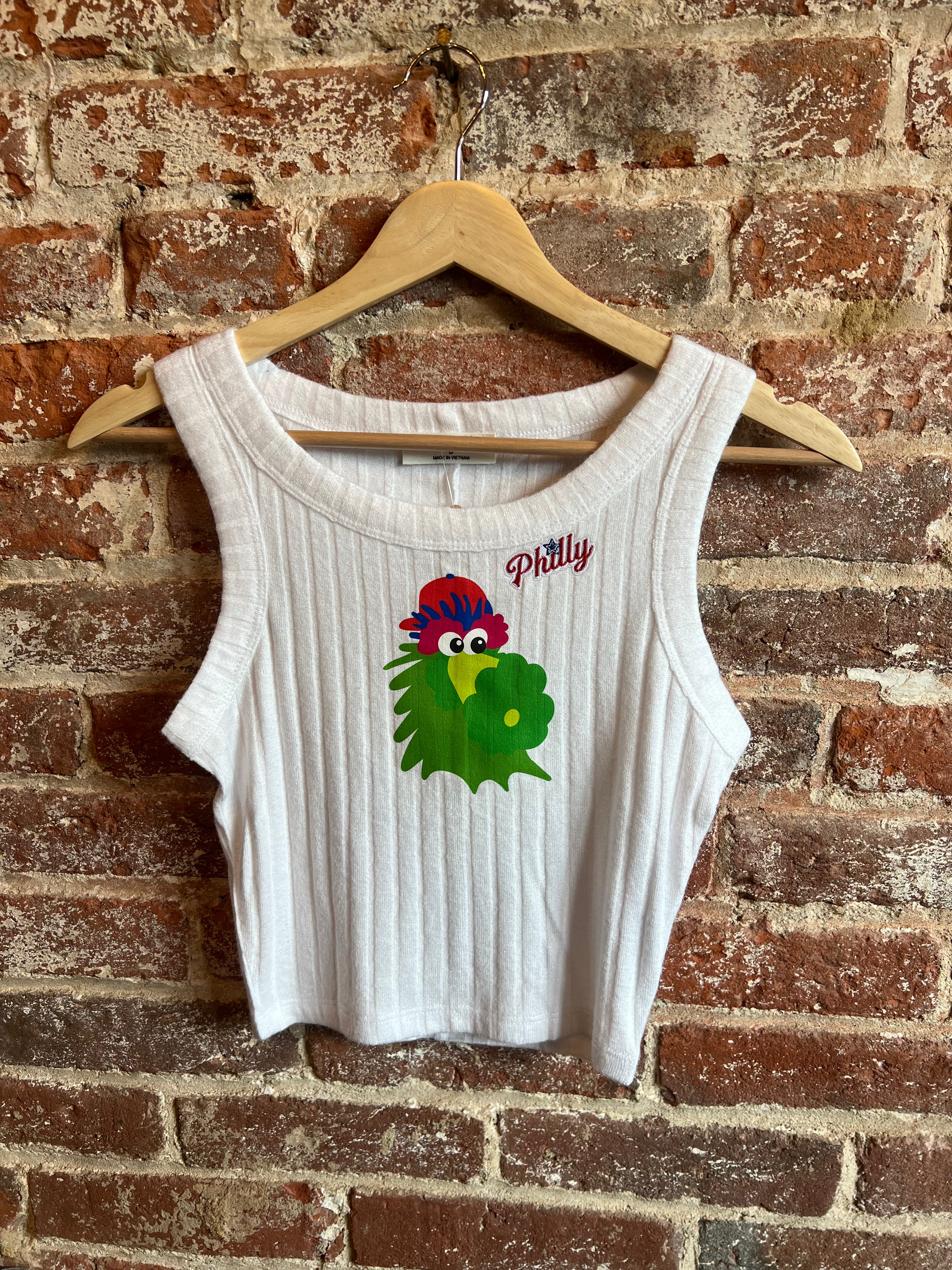 White tank top with a colorful parrot design and 'Pittily' text on a wooden hanger against a brick wall.
